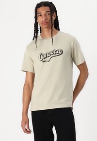 Camiseta de algodón color beige claro con un logo negro de "Converse" en cursiva en el pecho. Manga corta y corte estándar.