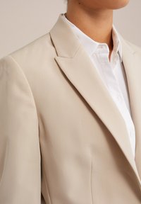 Beige sako s ostrým límcem, hladkým materiálem a strukturovaným střihem, vrstvené přes bílou košili s upraveným límcem a detaily na knoflících.