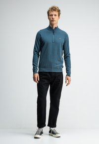 Blauer, langärmliger Pullover mit halbem Reißverschlusskragen, strukturiertem Stoff und einem Logo auf der Brust, kombiniert mit schwarzen Hosen und schwarzen Sneakern.