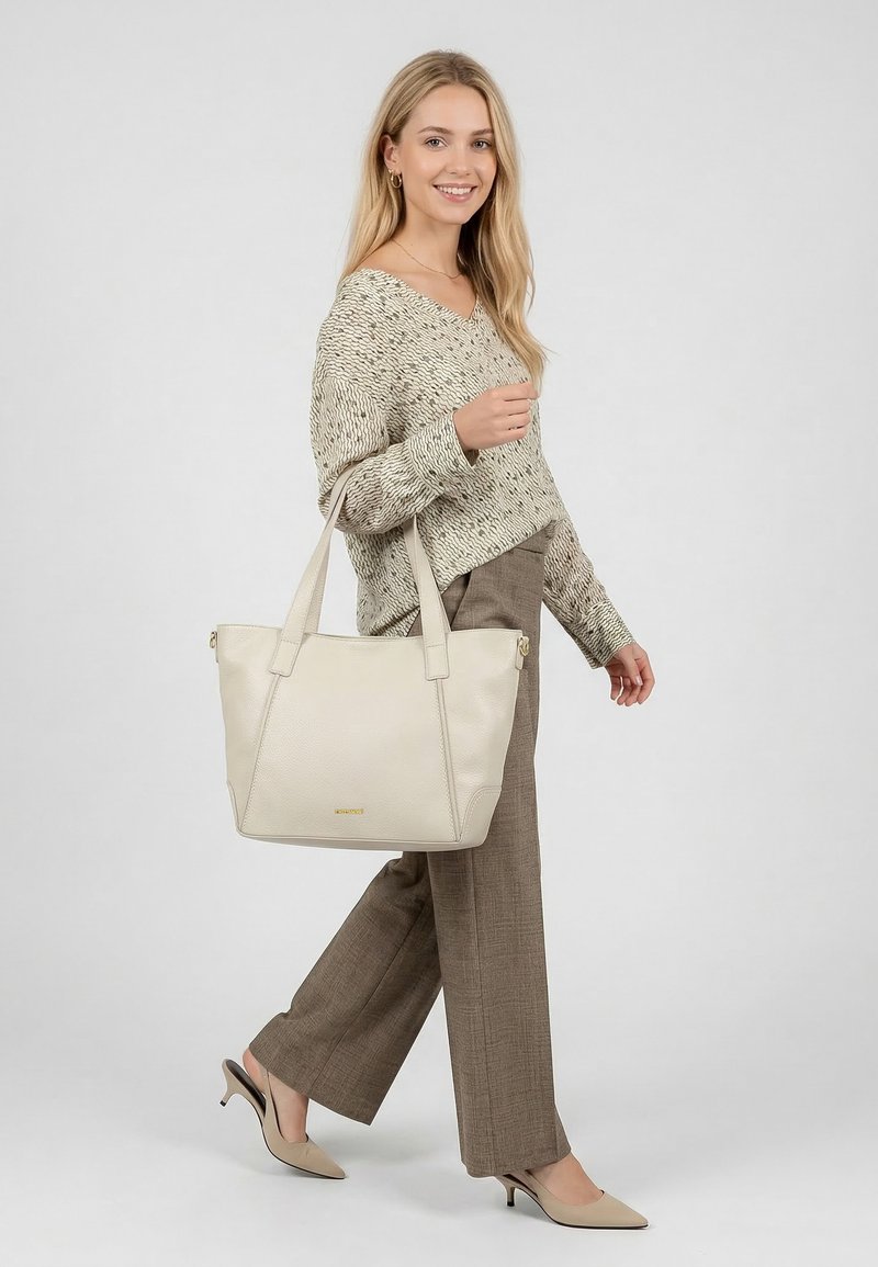 Junge Frau trägt einen beigefarbenen Pullover, braune weit geschnittene Hose und beige High Heels, mit einer großen offwhite Ledertasche über der Schulter.