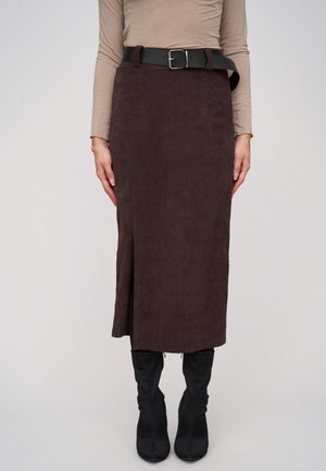 Femme portant une jupe midi en velours côtelé marron foncé avec une fente sur le côté, un haut beige à manches longues, une ceinture noire et des bottes noires hautes jusqu'aux genoux, debout devant un fond uni.