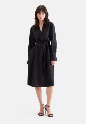 Kvinde med mørkt hår iført en sort midi-kjole med lange ærmer, taljebånd og sorte sandaler med høje hæle, stående på en ensfarvet baggrund.