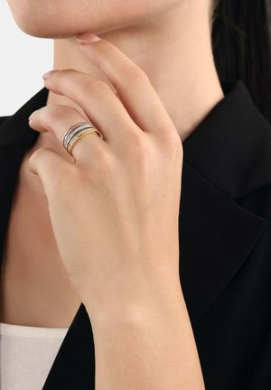 Anelli impilati con toni d'argento e oro, con fasce testurizzate e accenti lisci, indossati su una mano contro uno sfondo di blazer nero.