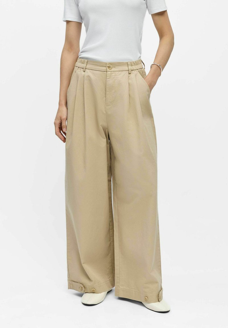 Femme portant un pantalon beige à jambes larges avec poignets boutonnés, un top blanc à manches courtes, des chaussures blanches et un bracelet argenté, une main dans la poche.