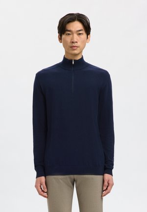 Jonge man die een marineblauwe pullover met lange mouwen en een korte rits aan de voorkant draagt, met beige broek, staand tegen een effen lichte achtergrond.