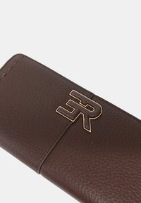 Portafoglio in pelle marrone con superficie strutturata, caratterizzato da un prominente emblema a forma di lettera dorata e un design di chiusura orizzontale.