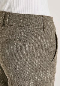 Close-up van een persoon die hoge, bruin getextureerde broeken draagt met riemlussen en insteekzakken, gecombineerd met een witte geribbelde top.