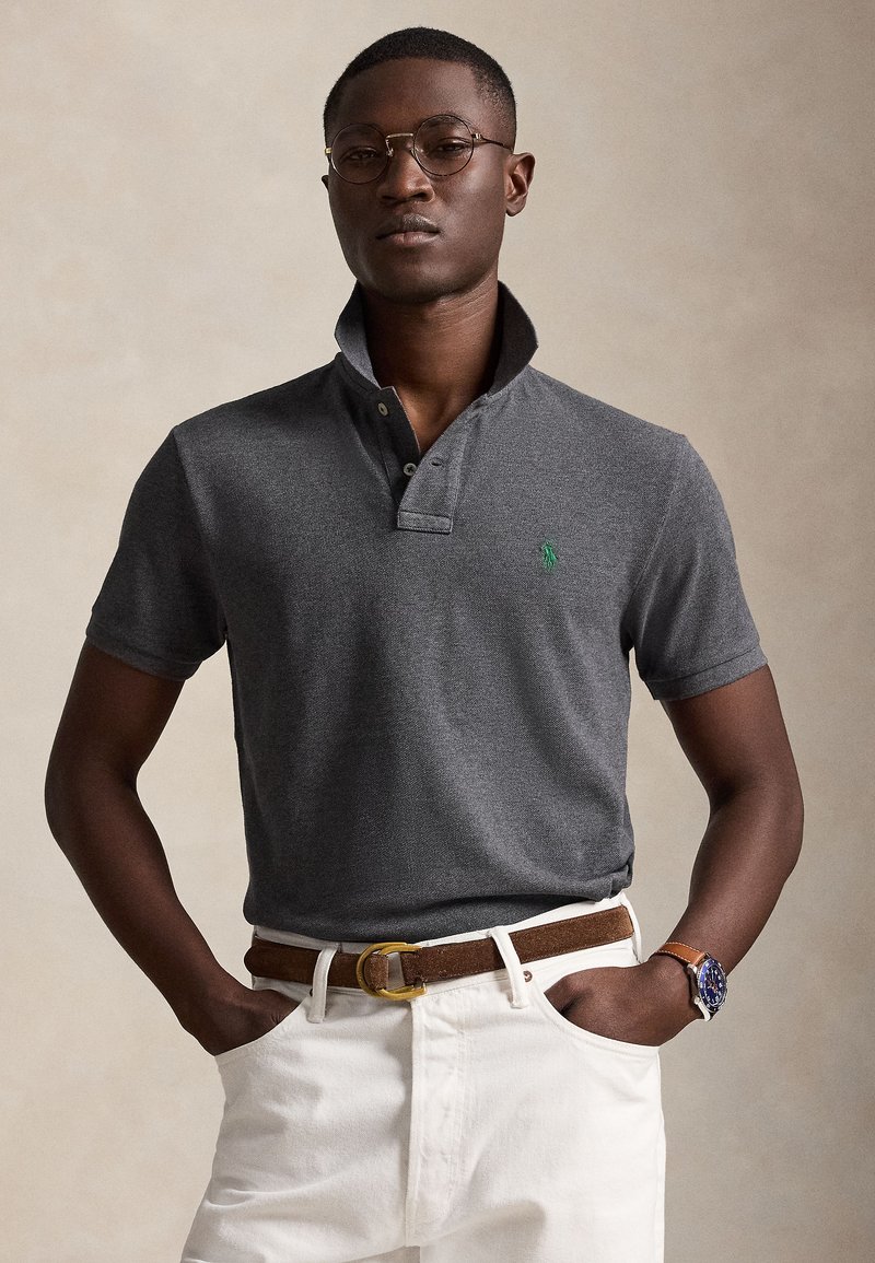 Homme portant des lunettes rondes, un polo gris foncé avec col relevé, un pantalon blanc, une ceinture marron et une montre-bracelet avec cadran bleu, les mains dans les poches.