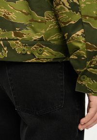 Μπουφάν από ύφασμα με καμουφλάζ σε αποχρώσεις ελιάς και καφέ, με δύο πράσινα κουμπιά στο κάτω μέρος, σε συνδυασμό με μαύρο τζιν παντελόνι.