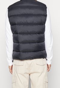 Man som bär en svart vadderad puffer väst över en vit långärmad skjorta och beiga cargobyxor, sedd bakifrån mot en enfärgad bakgrund.