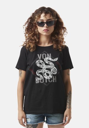 Femme aux cheveux bouclés portant des lunettes de soleil noires, un t-shirt noir avec des graphiques de serpent blancs et du texte, et un short en jean déchiré.