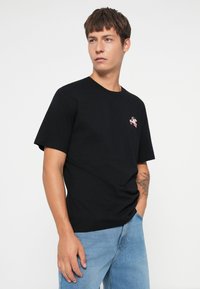 Black cotton t-shirt with short sleeves, round neckline, and a small colorful graphic on the left chest. Worn with light blue denim jeans.