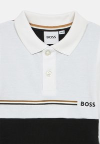Polo alb, negru și auriu, cu un gât ribetat și o deschidere cu trei butoane, prezintă dungi orizontale pe piept. Logo-ul brandului este afișat.