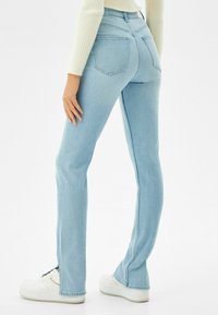 Vaaleansiniset farkut, joissa on slim fit -leikkaus, varustettu takataskuilla ja sauman yksityiskohdilla. Pukeutuneena valkoisiin lenkkareihin ja ribbattuun neuleeseen.