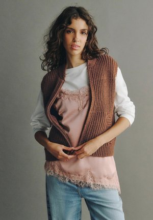 Jeune femme aux cheveux bouclés portant une chemise blanche à manches longues, un haut rose orné de dentelle, un gilet en tricot marron et un jean bleu clair, ajustant son gilet.