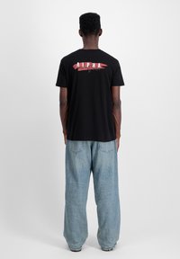 Sort bomulds t-shirt med korte ærmer, featuring "ALPHA" i rød og hvid på ryggen. Kombineret med løstsiddende, lyseblå denimbukser.