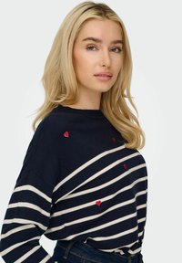 Maglione blu navy con strisce bianche orizzontali e ricamo di un piccolo cuore rosso. Tessuto morbido, scollatura rotonda, vestibilità rilassata.