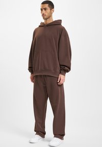 Ruskea fleece-huppari, jossa on etutasku ja joustavat hihansuut, yhdistettynä samanvärisiin ruskeisiin sweats-housuihin. Asusteessa on rento istuvuus.