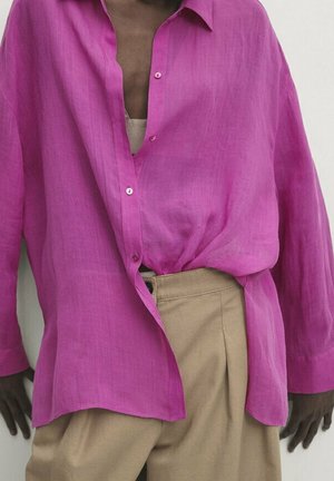Persona che indossa una camicia oversize rosa acceso con bottoni, infilata parzialmente in pantaloni beige a vita alta, in piedi davanti a uno sfondo semplice.