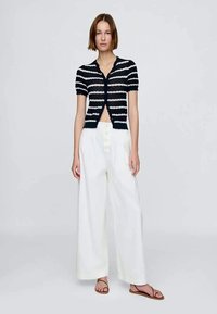 Mulher a usar uma blusa de manga curta às riscas preto e branco, calças largas brancas e sandálias castanhas, em frente a um fundo simples.