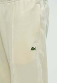 Μπεζ φόρμες από υφή ύφασμα, με λευκές ρίγες, πλαϊνές τσέπες και ένα πράσινο κεντημένο λογότυπο με κροκόδειλο στον αριστερό μηρό.