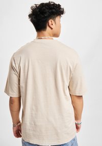 Ung person med krøllet mørkt hår iført en ensfarvet beige T-shirt og denimshorts, stående med ryggen til på en hvid baggrund.