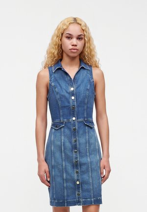 Jeune femme aux longs cheveux blonds bouclés portant une robe en denim bleue sans manches, boutonnée avec des poches, debout devant un fond uni.