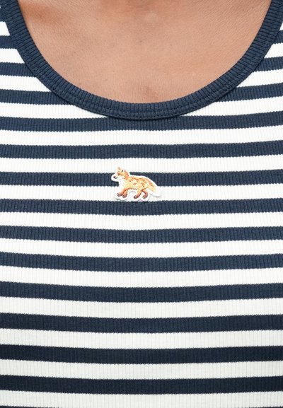 Tuvuma tuvā pie jūras un baltās svītrainās krekls ar mazu izšūtu oranžu lapsu emblemu zem apkakles.