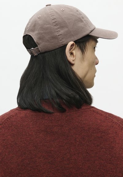 Casquette de baseball en coton marron avec une sangle ajustable à l'arrière, dotée d'une visière incurvée et d'un tissu texturé. Portée avec un pull en tricot rouge.