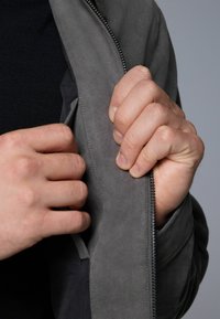Chaqueta de ante gris con una cremallera plateada. Una mano abre un bolsillo, mostrando un forro interior en contraste. Se aprecia una textura suave y un diseño ajustado.