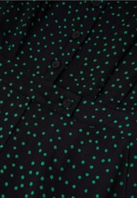 Tissu noir à pois verts irréguliers, avec des boutons noirs et un détail de ceinture sur un vêtement.