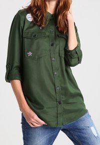 Camicia verde a maniche lunghe realizzata in tessuto morbido. Presenta due tasche sul petto, bottoni neri e toppe decorative sul petto.