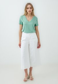T-shirt verde chiaro a V con maniche corte, abbinato a pantaloni bianchi a gamba larga e sandali bianchi con cinturini, su uno sfondo neutro.