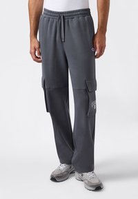 Mann trägt dunkelgraue Cargo-Sweatpants mit elastischem Bund und wei�ßen Sneakern, steht vor einem schlichten weißen Hintergrund.