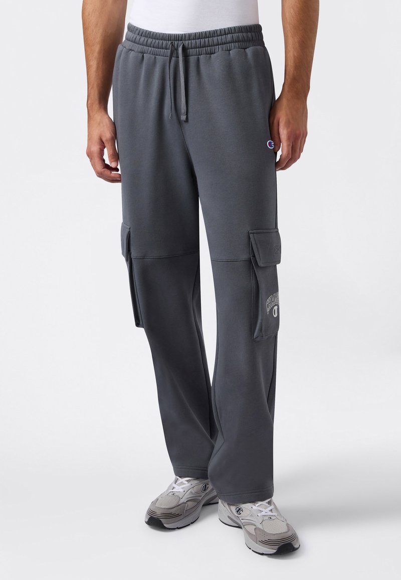 Mann trägt dunkelgraue Cargo-Sweatpants mit elastischem Bund und weißen Sneakern, steht vor einem schlichten weißen Hintergrund.