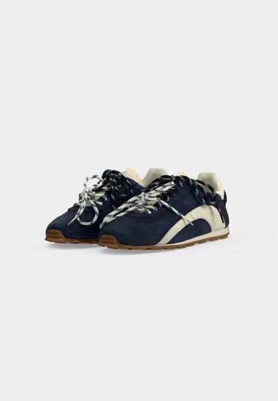 Paire de baskets bleu marine et blanches avec des lacets à motifs et des semelles marron, vues de face sur un fond uni.