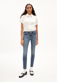 Witte short-sleeve button-up shirt, gecombineerd met strakke blauwe jeans. Het model draagt zwarte instappers met witte sokken. Eenvoudig, strak ontwerp.