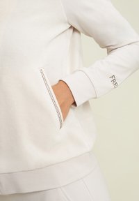 Felpa beige chiaro realizzata in tessuto morbido, dotata di tasca laterale con dettagli metallici e polsino con un motivo sottile.