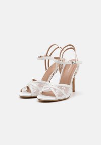 Sandales blanches à talons hauts avec un design à brides croisées, des détails en dentelle et des lanières autour de la cheville. Elles sont dotées d'une semelle lisse et d'un talon aiguille élancé.