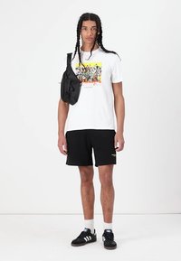 White graphic t-shirt with colorful print, black shorts, black shoulder bag, and black sneakers with white accents, standing on a plain background.