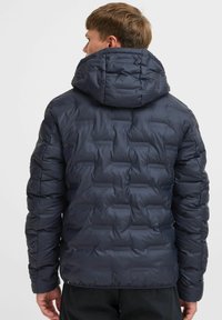 Mørkeblå pufferjakke med et struktureret design, der har hætte og kviltede paneler for tekstur. Fremstillet af glat, vandafvisende materiale.