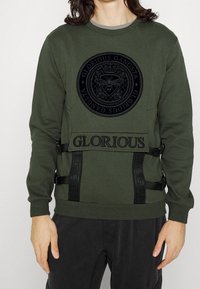 Persoon in een olijfgroene sweatshirt met zwart bij-embleem en "Glorious Gangsta" tekst, gecombineerd met zwarte broek, staand tegen een witte achtergrond.