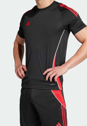 Mand iført sort atletisk t-shirt og shorts med røde og hvide detaljer, hænderne på hoften, overskroppen og taljen synlige.