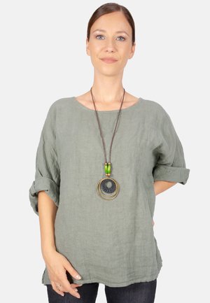 Grüne Leinenbluse mit aufgerollten Ärmeln und runder Ausschnitt, kombiniert mit einer mehrlagigen Anhängerkette mit grünen und goldenen Akzenten.