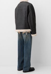 Personne portant une veste en shearling noir avec des bordures en fourrure beige, un jean bleu ample avec des poches arrière uniques en surpiqûre, et des mocassins noirs.