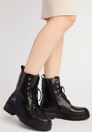 Bottes de combat en cuir noir à lacets avec semelles épaisses, portées par une personne portant une jupe beige sur un fond uni.