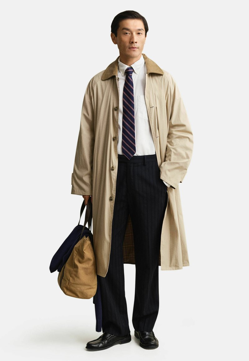 Mann in beigem Trenchcoat, marineblau gestreifter Krawatte, weißem Hemd, schwarzen Nadelstreifenhosen, hält eine beige-marineblaue Tasche, steht vor weißem Hintergrund.