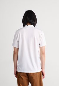 Bílá polo košile s krátkým rukávem, hladkým povrchem a klasickým límečkem, s čistým, minimalistickým designem a rovnými okraji na zadní straně.