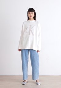 Bela pletena jopica z žametasto teksturo in velikim bež logotipom "cK", v kombinaciji s svetlo modrimi širokimi jeans hlačami in srebrnimi balerinkami.