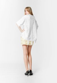 Camisa blanca de gran tamaño con detalles en la espalda, combinada con una falda amarilla texturizada con pliegues y zapatos de tacón negros.